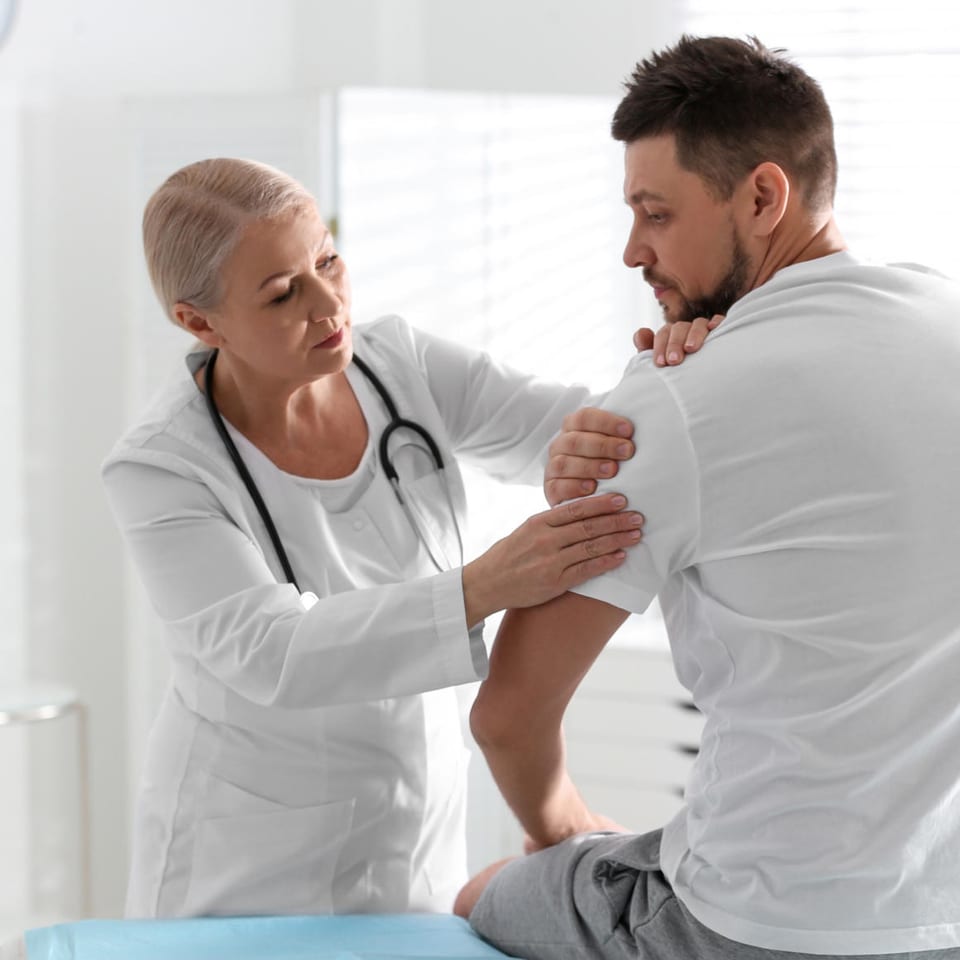 A female doctor in a white coat examining a male patient's shoulder in a clinic.