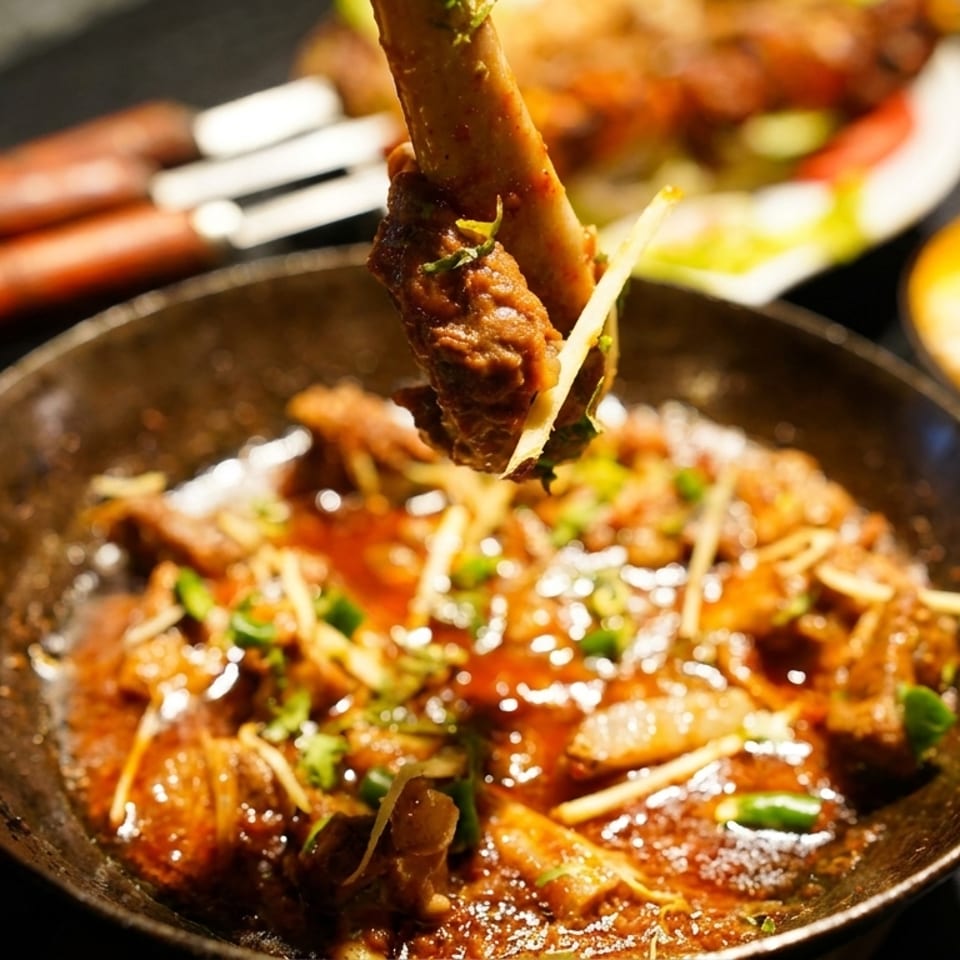 A hand holding a lamb shank over a karahi of flavorful curry with ginger and cilantro.
