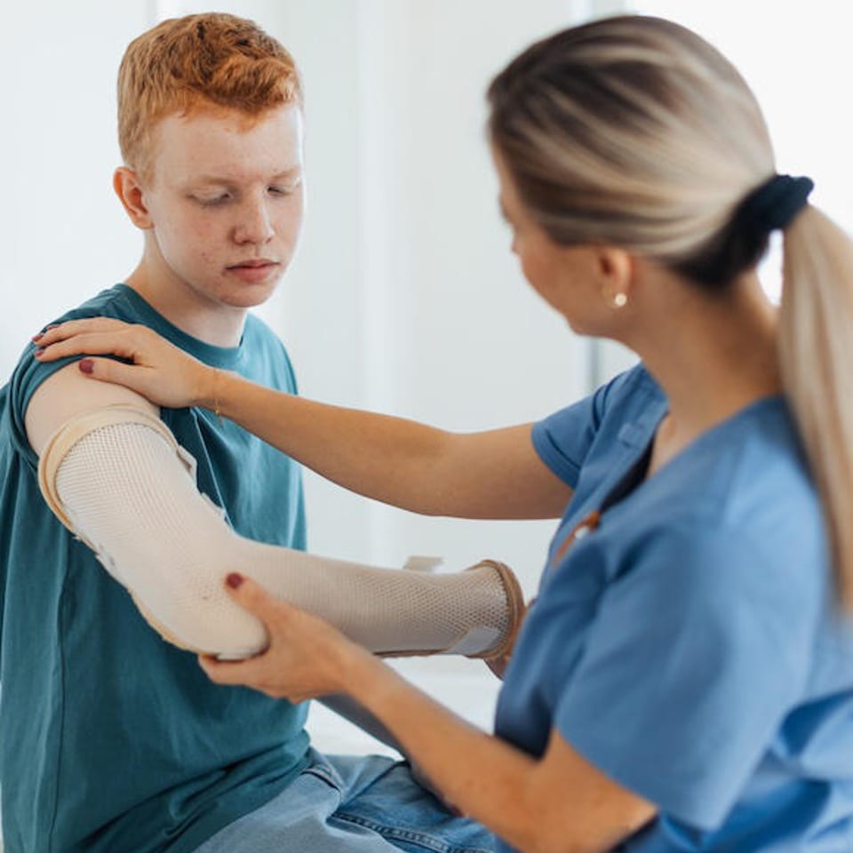 A nurse in blue scrubs gently adjusts a young man's arm cast.